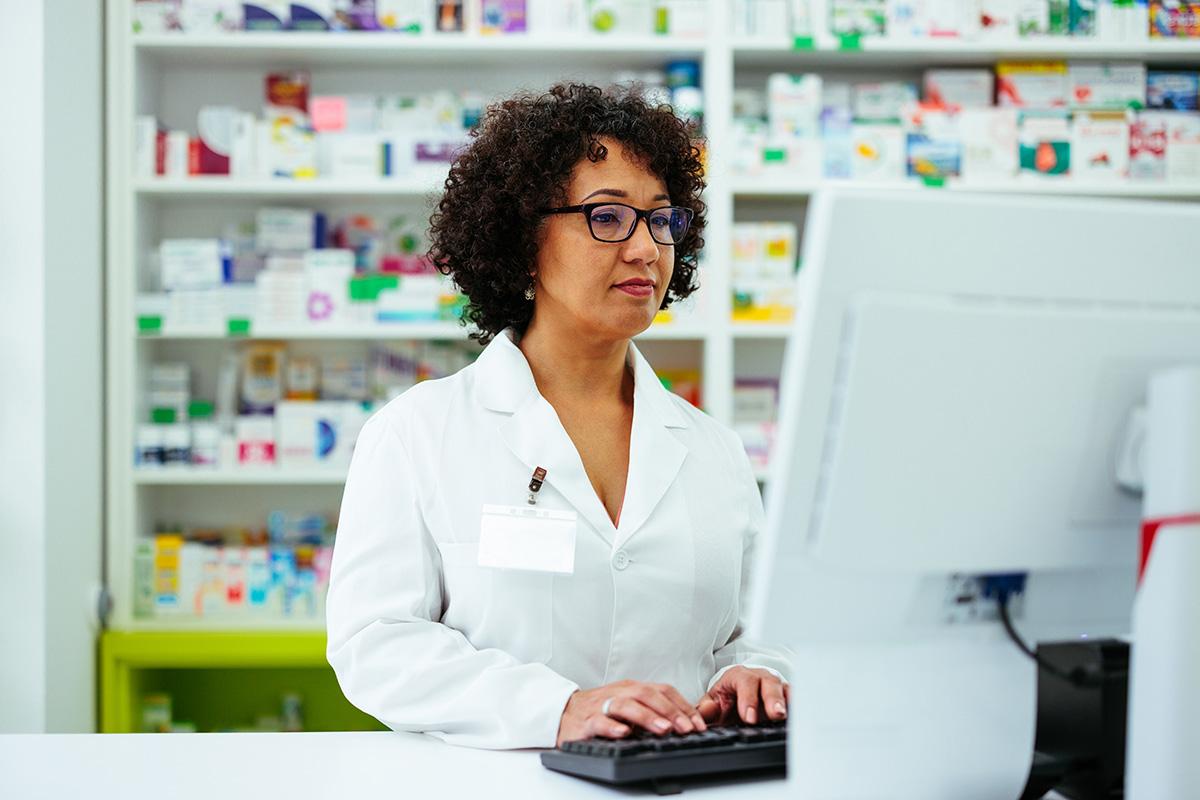 pharmacy technician working on a computer with medicines on shelves in background